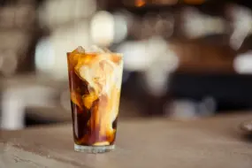The cream is swirling through the dark coffee, creating a marbled effect of white and brown. The drink is served in a clear glass with ice cubes, and it’s sitting on a counter or table with a blurred background, likely in a café or coffee shop setting. The lighting is warm and inviting, adding to the cozy vibe.
