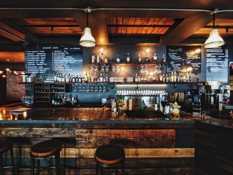 This image shows a stylish, rustic bar with a warm, inviting atmosphere. The bar counter is made of polished wood with rich tones, and three stools are neatly placed in front. Behind the bar, shelves are stocked with various bottles of liquor, glassware, and bar tools. Soft pendant lights hang from the ceiling, illuminating the space and highlighting the chalkboard menus that list drink options. The overall vibe is cozy and perfect for social gatherings or casual drinks.